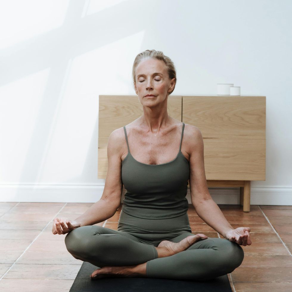 Eine Frau mit grauen Haaren sitzt in Yogakleidung auf einer Yogamatte im Lotussitz.