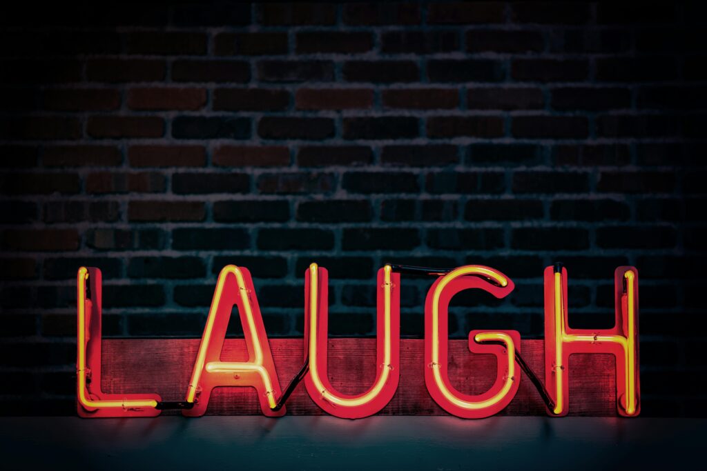 Wie sich Humor auf unsere Psychoe auswirkt: Auf dem Bild ist eine Leuchtschrift mit dem Schriftzug "Laugh" zu sehen, im Hintergrund eine dunkle Ziegelmauer