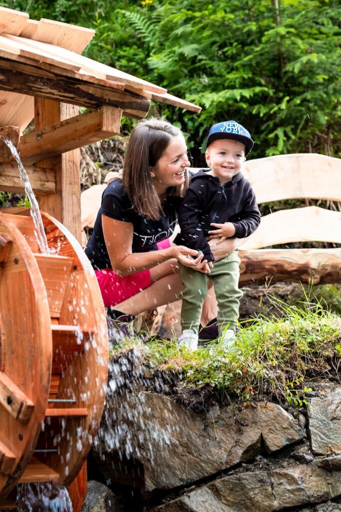 Wildkogel-Arena Neukirchen & Bramberg: Eine Mutter mit Kind, lachend, vor einer Holzmühle, die mit Wasser angetrieben wird