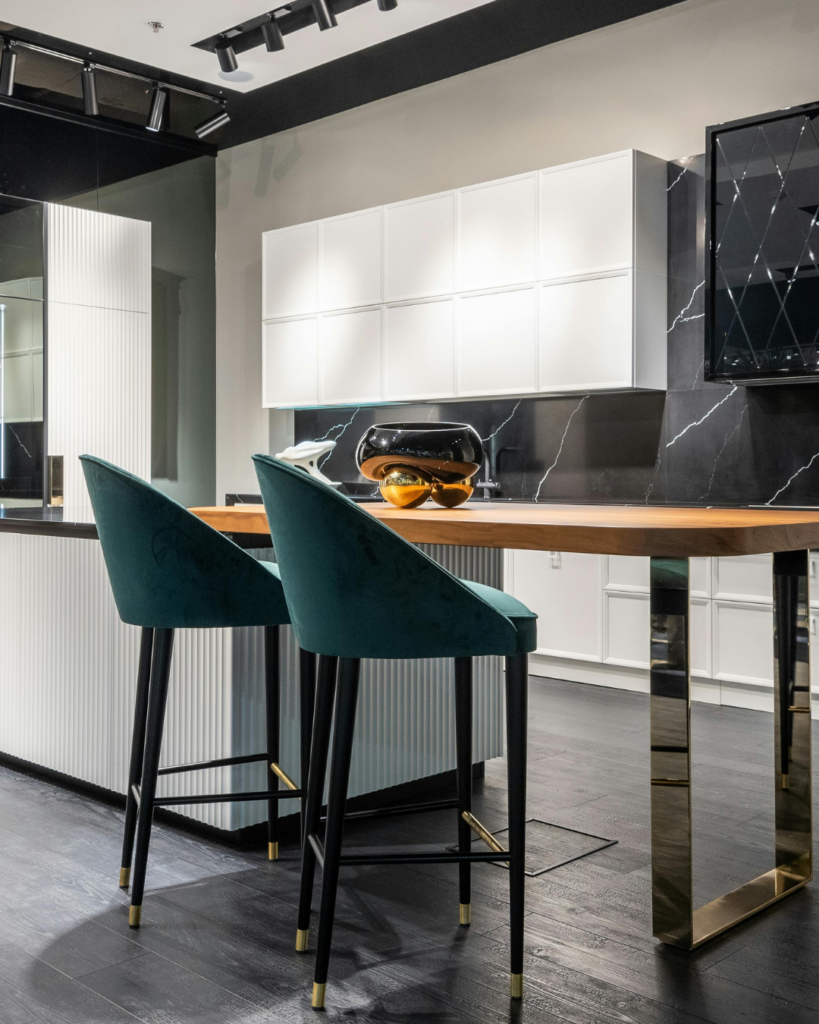 Eine stylische Küche mit zwei grünen Stühlen im Vordergrund, ein Holztisch mit Füßen aus Eisen und an der Wand ein weißer Hängeschrank.