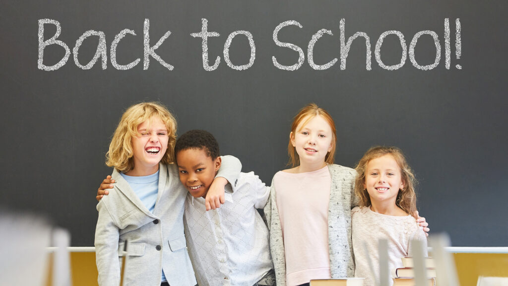 Schulbeginn: Schulkinder vor der Tafel mit dem Schriftzug ‚Zurück zur Schule‘.
