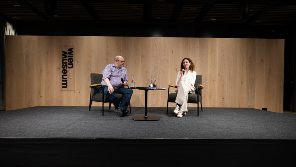 Judith Kohlenberger: Zwei Personen sitzen auf einer Bühne im Wien Museum und führen ein Gespräch, beide halten Mikrofone.