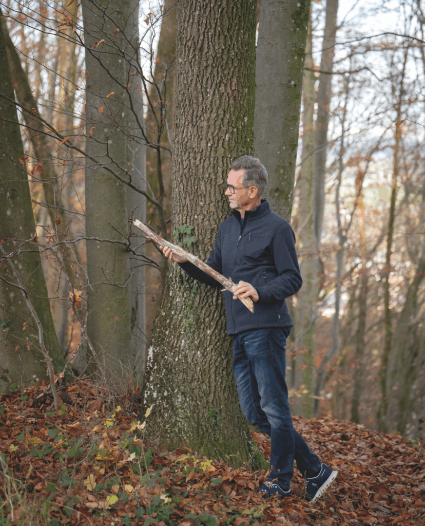 Im Bild zu sehen: Autor Gerald Ziegler im Wald, wie er an einem Baum anlehnt und einen Ast in der Hand hält