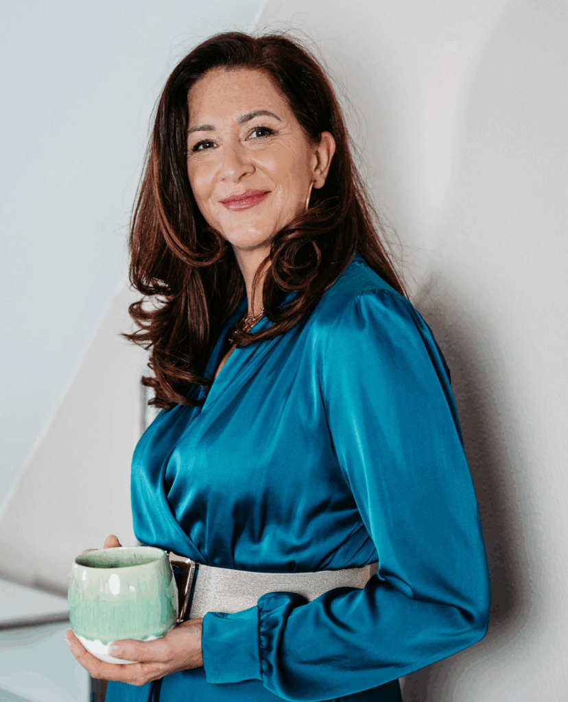 Autorin Ute Zischinsky in blauem Satin-Oberteil, hält eine Tasse in der Hand und lächelt in die Kamera