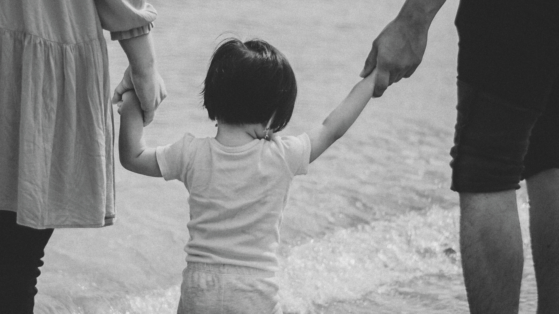 Ein Kind wird links und rechts an der Hand gehalten und geht einen Strand entlang