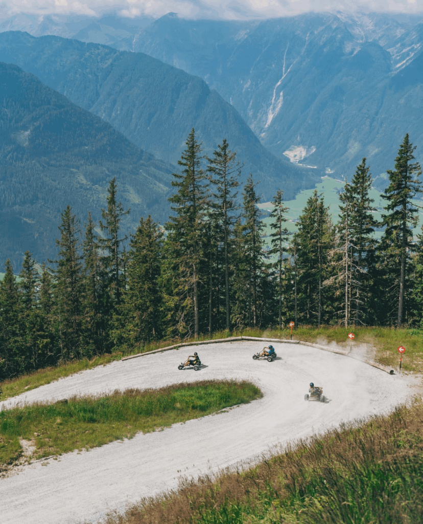 Ein wunderschönes Panorama in der Wildkogel-Arena mit mehreren Personen, die auf Mountaincarts eine kurvenreiche Schotterstrecke durch einen dichten Wald hinunterfahren, umgeben von beeindruckenden Bergen im Hintergrund.