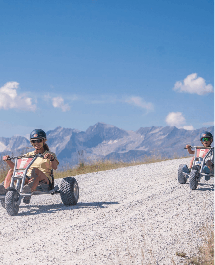 Ein wunderschönes Panorama in der Wildkogel-Arena mit mehreren Personen, die auf Mountaincarts eine kurvenreiche Schotterstrecke durch einen dichten Wald hinunterfahren, umgeben von beeindruckenden Bergen im Hintergrund.