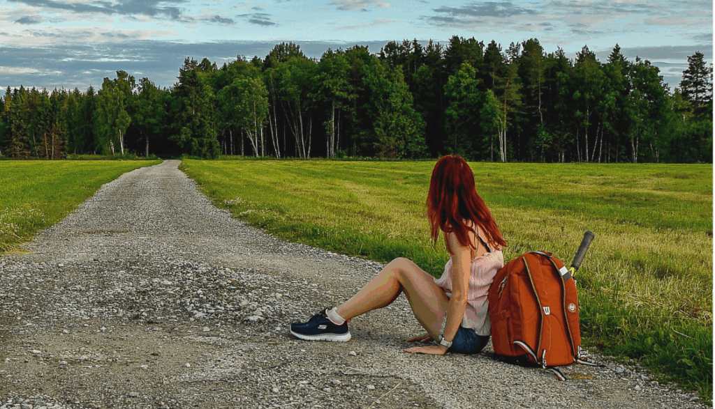 Mädchen mit roten Haaren sitzt am Boden mit Rucksack, blickt in die Wanderlandschaft