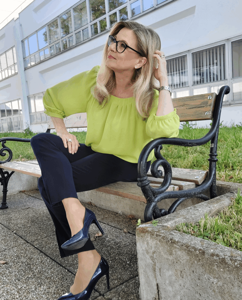 Christa Kummer auf einer Bank, lachend zur Seite schauend im grünen Oberteil und schwarzer Hose, schwarze Stöckelschuhe