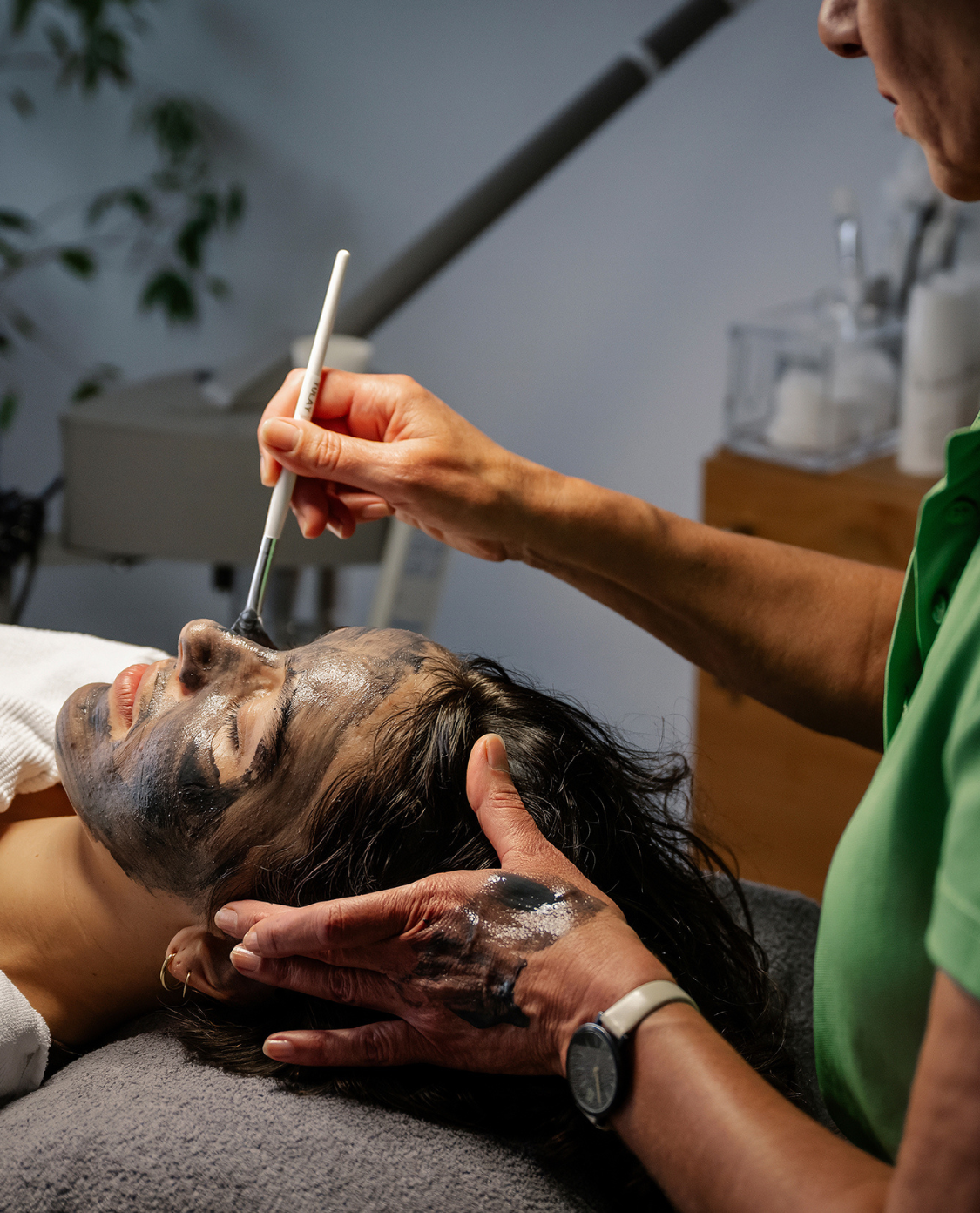Eine Frau, die eine Gesichtsbehandlung bekommt, sie liegt und eine andere Frau trägt ihr mit Pinsel eine dunkle Maske auf