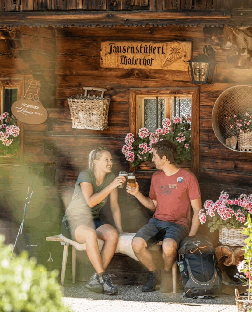 Ein Pärchen, das vor einer Almhütte sitzt und mit Bier anstößt.