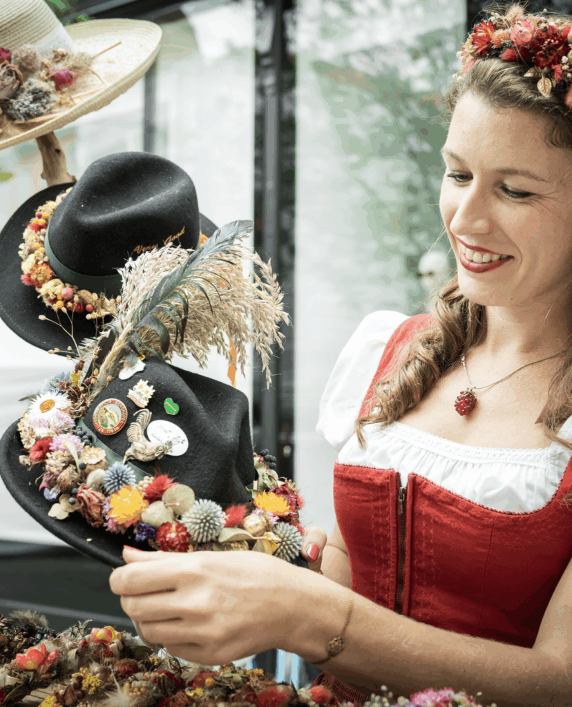 Eine Frau in Tracht, die sich festlich geschmückte Trachtenhüte ansieht. Einen davon hält sie in der Hand.
